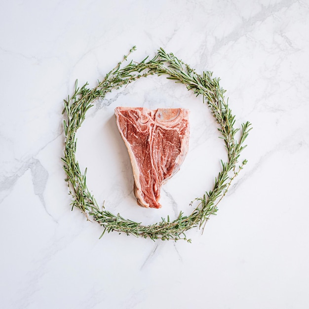 Raw tbone steak in the center of a Christmas wreath of rosemary thyme cherry tomatoes and bell peppers on a white marble surface christmas dinner concept