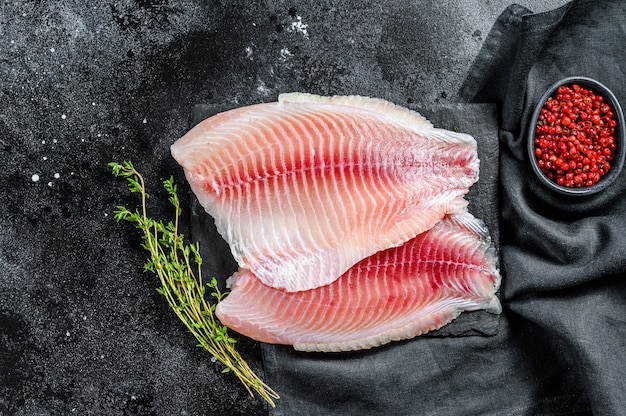 Raw telapia fillet on a chopping Board with thyme and pink pepper. Black background. Top view