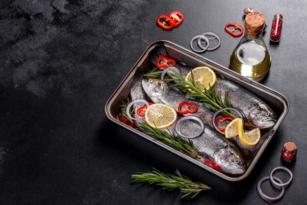 Raw trout lie on a black stone chopping board on a dark concrete table. Nearby are: tomato, rosemary, lemon, pepper, onion, salt