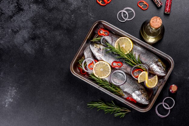 Raw trout lie on a black stone chopping board on a dark concrete table. Nearby are: tomato, rosemary, lemon, pepper, onion, salt