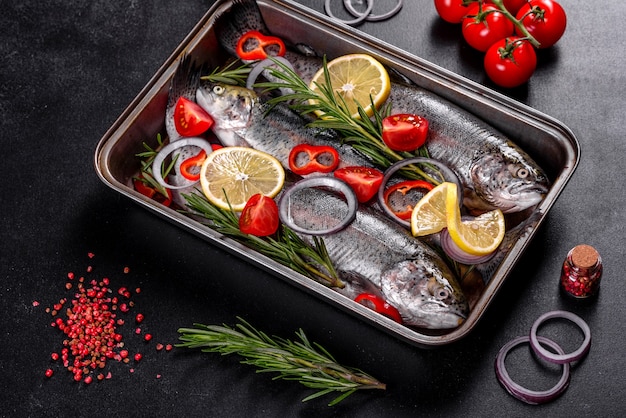 Raw trout lie on a black stone chopping board on a dark concrete table. Nearby are: tomato, rosemary, lemon, pepper, onion, salt