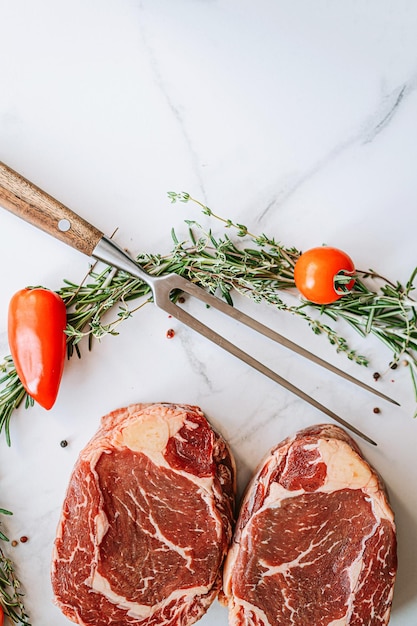 Raw two ribeye steaks in the center of a Christmas wreath of rosemary thyme cherry tomatoes and bell peppers on a white marble surface christmas dinner concept for two