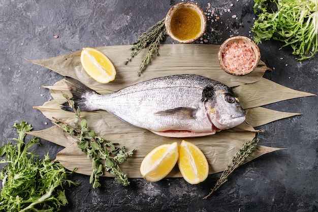 Raw uncooked fresh gutted sea bream or dorado fish served with lemon, green salad, rosemary, olive oil and pink salt on bamboo leaves over dark texture surface. Flat lay, copy space.