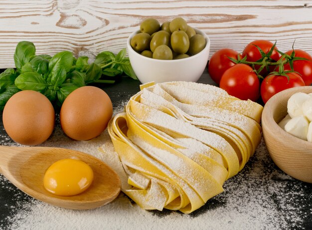 Raw yellow italian pasta pappardelle, fettuccine or tagliatelle close up. Egg homemade noodles cooking process, long rolled macaroni or uncooked spaghetti with olives, tomatoes, basil