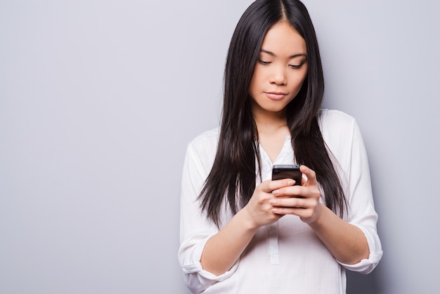 Reading new messages. Beautiful young Asian woman holding mobile phone while standing against grey background