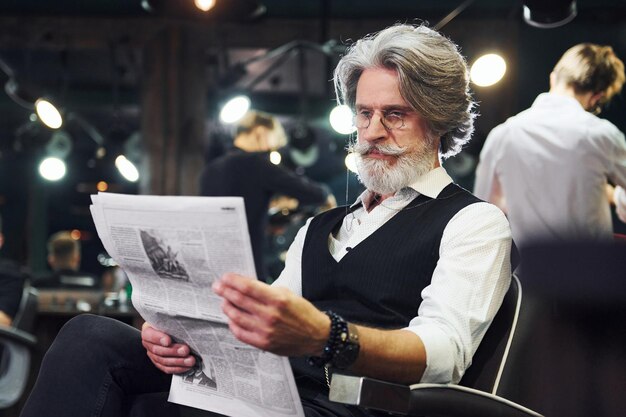 Reading newspaper Stylish modern senior man with gray hair and beard is indoors