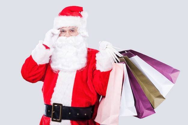 Ready for Christmas sales? Happy Santa Claus holding shopping bags and adjusting his eyeglasses while standing against grey background