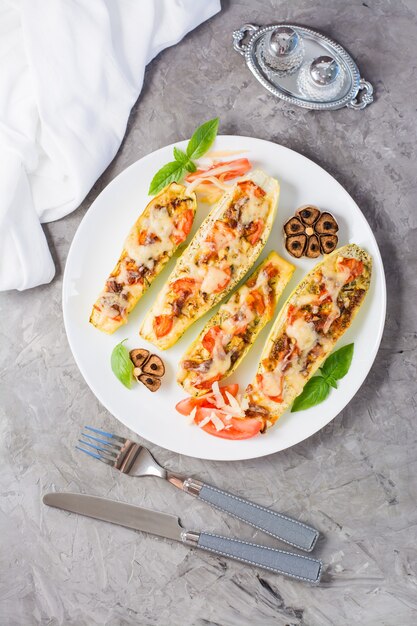 Ready-to-eat baked zucchini halves stuffed with cheese and tomato and basil leaves on a plate on the table. Vegetable menu, healthy food. Vertical and top view