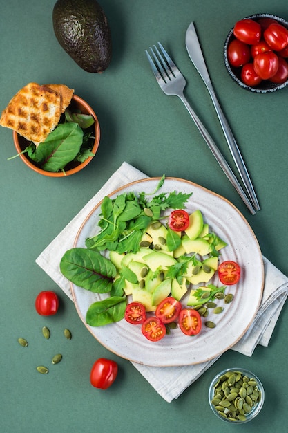 Ready-to-eat salad with avocado, tomatoes, arugula leaves, mizuna, chard and pumpkin seeds on a plate and cutlery on a green table. Vegetarian vitamin food. Top and vertical view