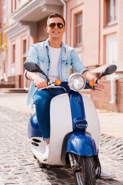 Ready to ride. Handsome young man riding scooter along the street and smiling