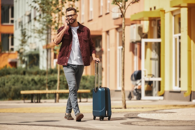 Ready for trip cheerful bearded man in casual wear and eyeglasses pulling his luggage and