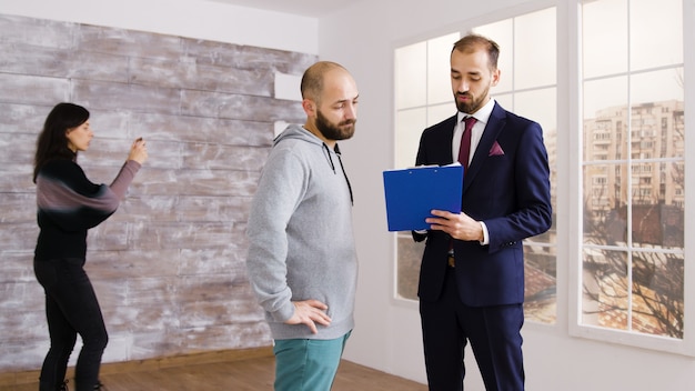 Photo real estate agent in business suit describes apartment to client in empty property while woman is taking photos in the background.