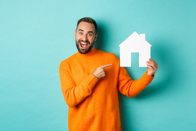 Real estate concept. Happy man pointing finger at paper home maket and smiling, standing over light blue background.