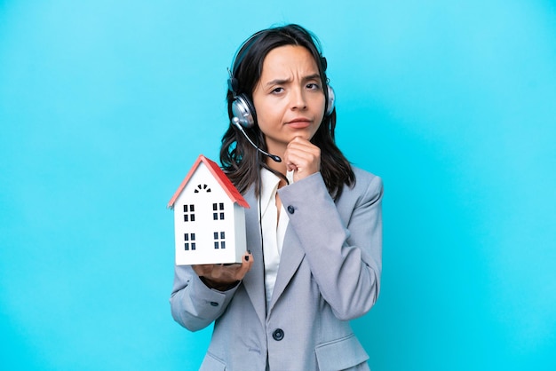 Real estate hispanic agent holding a toy house isolated on blue background thinking