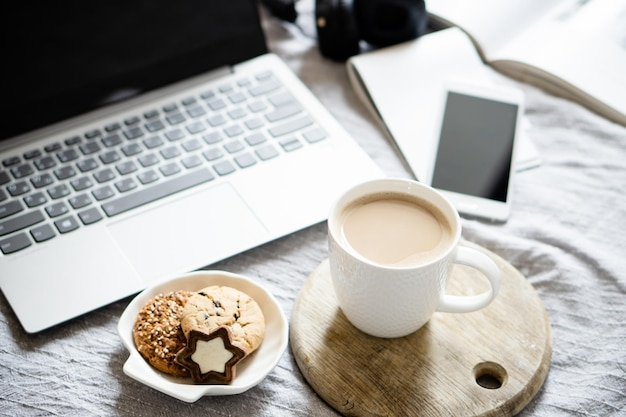 Real home workplace laptop with cup of coffee with cookies on bed with cozy blanket