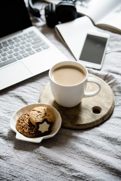 Real home workplace laptop with cup of coffee with cookies on bed with cozy blanket