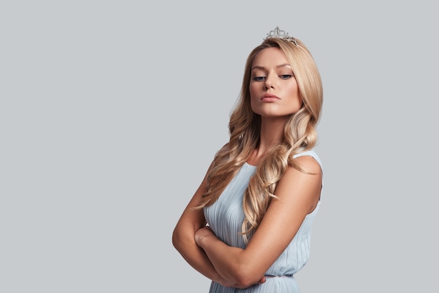 Real queen. Prideful young woman in crown keeping arms crossed while standing against grey background