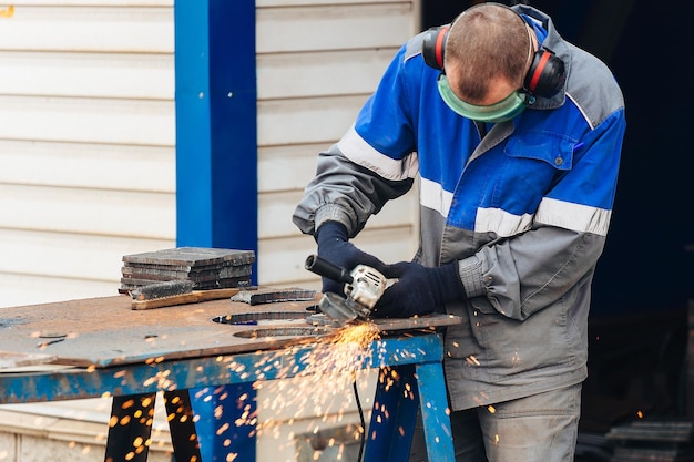 Real worker in working clothes grinds metal surface and sparks fly person at work