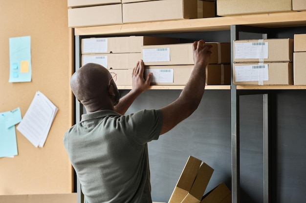 Rear view of african man putting finishes parcels in cardboard boxes on shelf during his work in war