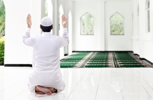 Rear view of Asian Muslim man sitting while raised hands and praying on the mosque