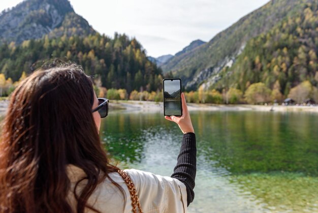 Rear view of beautiful young woman using mobile phone taking photos of beautiful lake