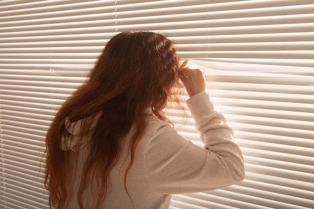 Rear view of beautiful young woman with long hair peeks through hole in the window blinds and looks