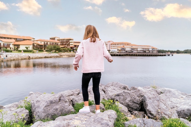 Rear view of a blonde Caucasian girl walking among stones in a village lake