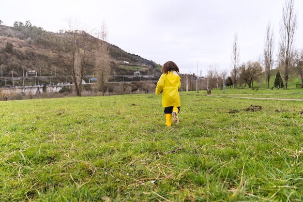 Rear view of a boy in a raincoat and yellow rain boots running across a park lawn