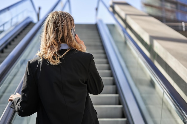 Rear view of businesswoman in suit standing on escalator and talking on mobile phone