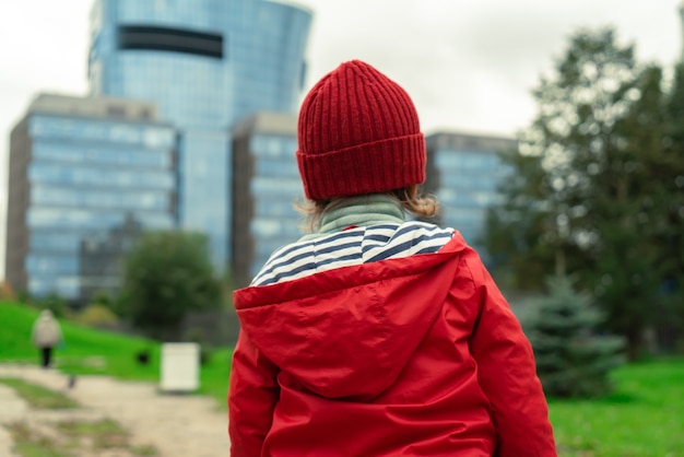 Rear view of a child in a red jacket and hat standing on the background of a modern cityscape view