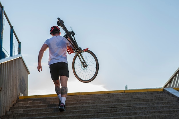 Rear view full length shot of a male cyclist walking up the stairs outdoors in the summer, carrying his bike, copy space