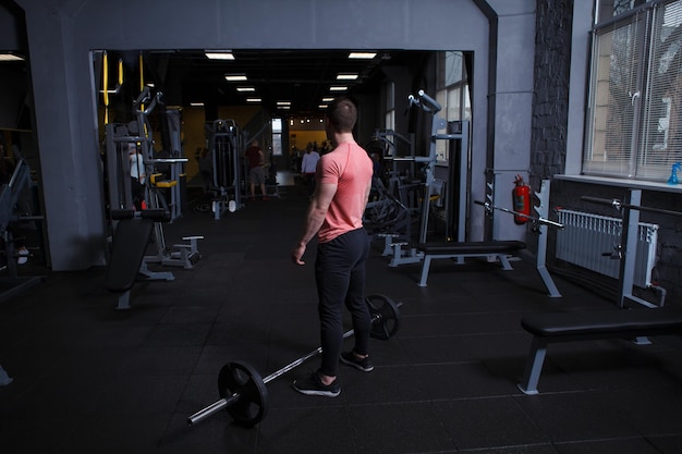 Rear view full length shot of a muscular sportsman standing in front of a barbell at the gym