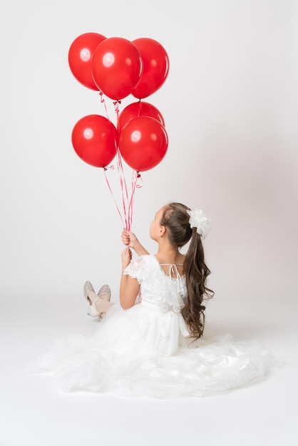 Rear view of girl in white dress holding lot of red balloons in hand sitting on white background