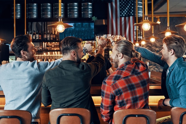 Rear view of happy young men in casual clothing toasting each other with beer and laughing while sitting in the pub