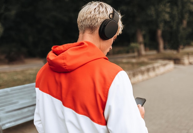 Rear view image handsome young blond man texting messages while listening to his favorite music from wireless headphone using smart phone walking outdoor in the city park