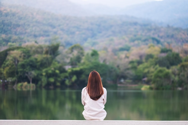 Rear view image of a woman sitting alone by the lake looking at the mountains with green nature background