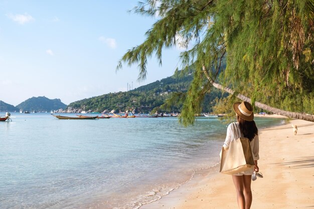 Rear view image of a woman with hat and bag strolling on the beach with blue sky background