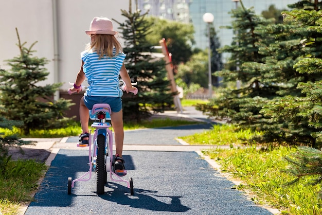 Rear view of little child in summer hat riding bike outdoors in city park