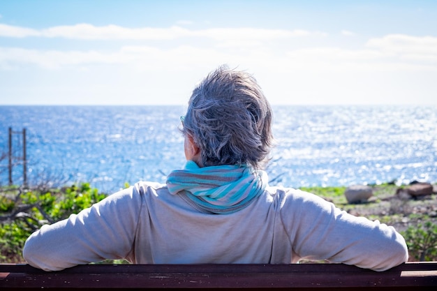 Rear view of mature grey-haired woman relaxing in front to the sea, sitting on a bench in a windy day. Senior woman enjoying retirement looking at horizon over water