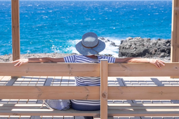 Rear view of mature woman dressed in blue admiring the sea looking at horizon over water Mature lady enjoying relaxation and vacation sitting on a wooden bench