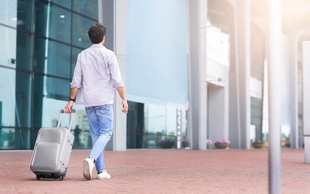 Rear view portrait of man walking outside of airport with suitcase