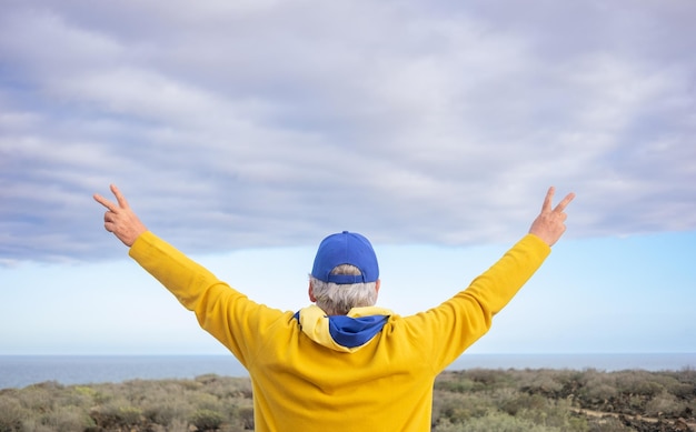 Rear view of senior man standing in front of the sea with the Ukrainian flag around his neck with outstretched arms and victory sign Message of freedom for the Ukrainian people