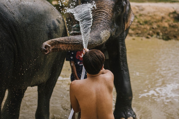 Photo rear view of shirtless man washing elephant with water pipe while standing in lake