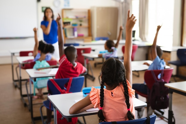 Rear view of students raising their hands while sitting on their desk in the class at school