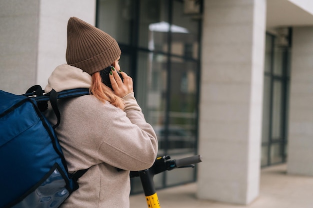 Rear view of unrecognizable female courier talking on phone with food restaurant or with customer delivering order on electric scooter