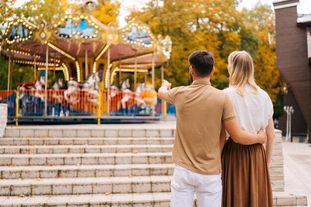 Rear view of unrecognizable loving young couple standing embracing watching rotation of carousel in amusement park