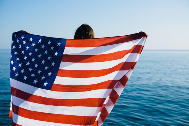 Rear view of unrecognizable young woman holding american waving flag at coastline against of sunny bright sea