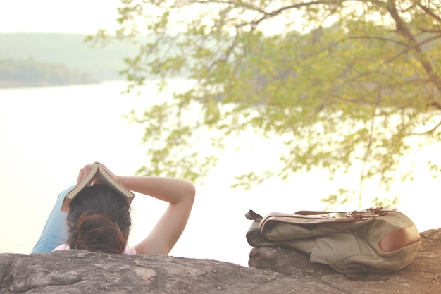 Photo rear view of woman sitting with book and bag by rock at riverbank during sunset