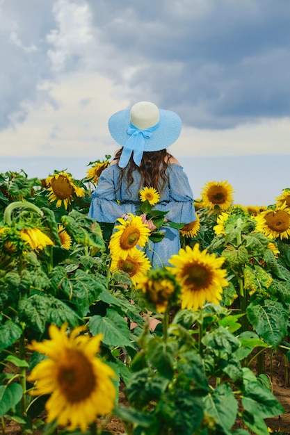 Rear view of a woman stands in sunflower field with her hands behind her back and holds sunflower