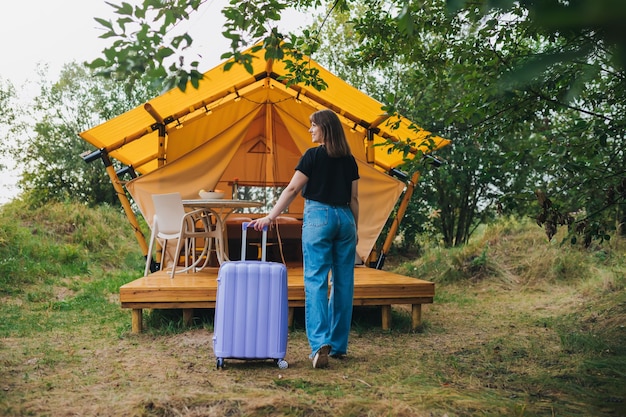 Rear view of woman traveler with luggage moving towards bedroom of cozy glamping house Luxury camping tent for outdoor summer holiday and vacation Lifestyle concept